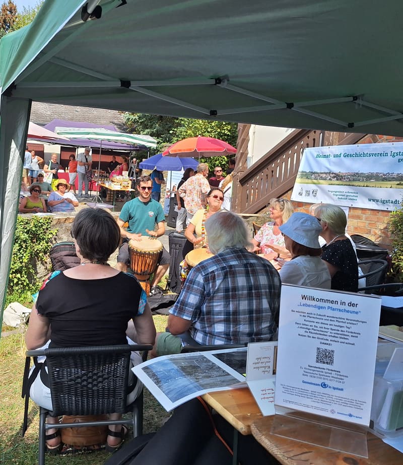 Infostand und Trommelgruppe des Förderkreises Igstadt an der Pfarrscheune Blick aus einem Pavillon auf einen Mann mit Trommel und Menschen an Tischen im Pfarrhof. Rechts ist ein Infostand des Förderkreises Igstadt mit Aufsteller und einem Banner des Heimat- und Geschichtsvereins Igstadt zu sehen